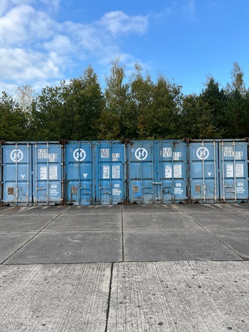 Blue shipping containers lined up outdoors, surrounded by trees under a cloudy sky.